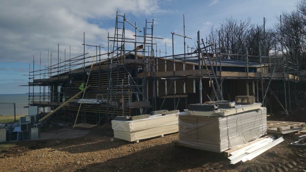 Scaffolding surrounds the exterior of Whitburn Coastal Conservation Centre, a small building under construction on the edge of the car park at Souter Lighthouse. Piles of building materials sit in front of it, the sea can be seen beyond.